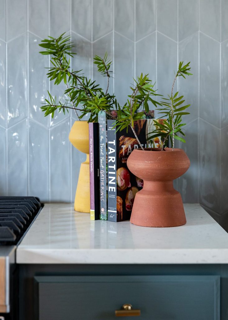 Handmade tile backsplash laid with a vertical stack. Modern subway tile. Cookbook styling with colorful vases. Marble and quartz countertops with blue cabinets and brass accents and hardware. 